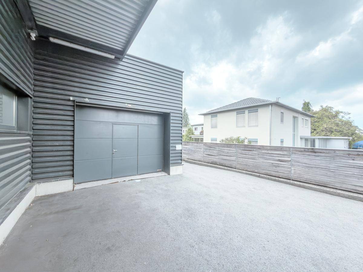 Außenansicht eines modernen Industriegebäudes mit Garagentor und Zufahrt, im Hintergrund ein Wohnhaus.