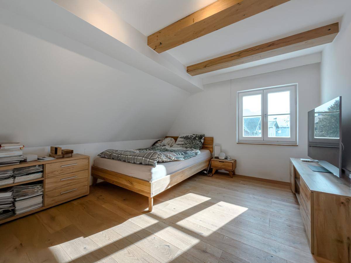 Gemütliches Schlafzimmer mit Holzbalken, Holzboden und viel Tageslicht durch das Fenster.