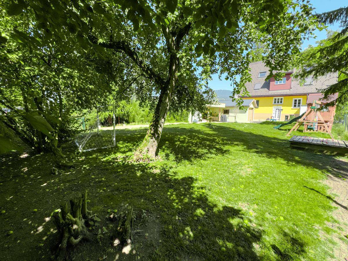 Idyllischer Gartenbereich unter einem großen Baum mit Rasenfläche und Blick auf das Haus und den Spielplatz.