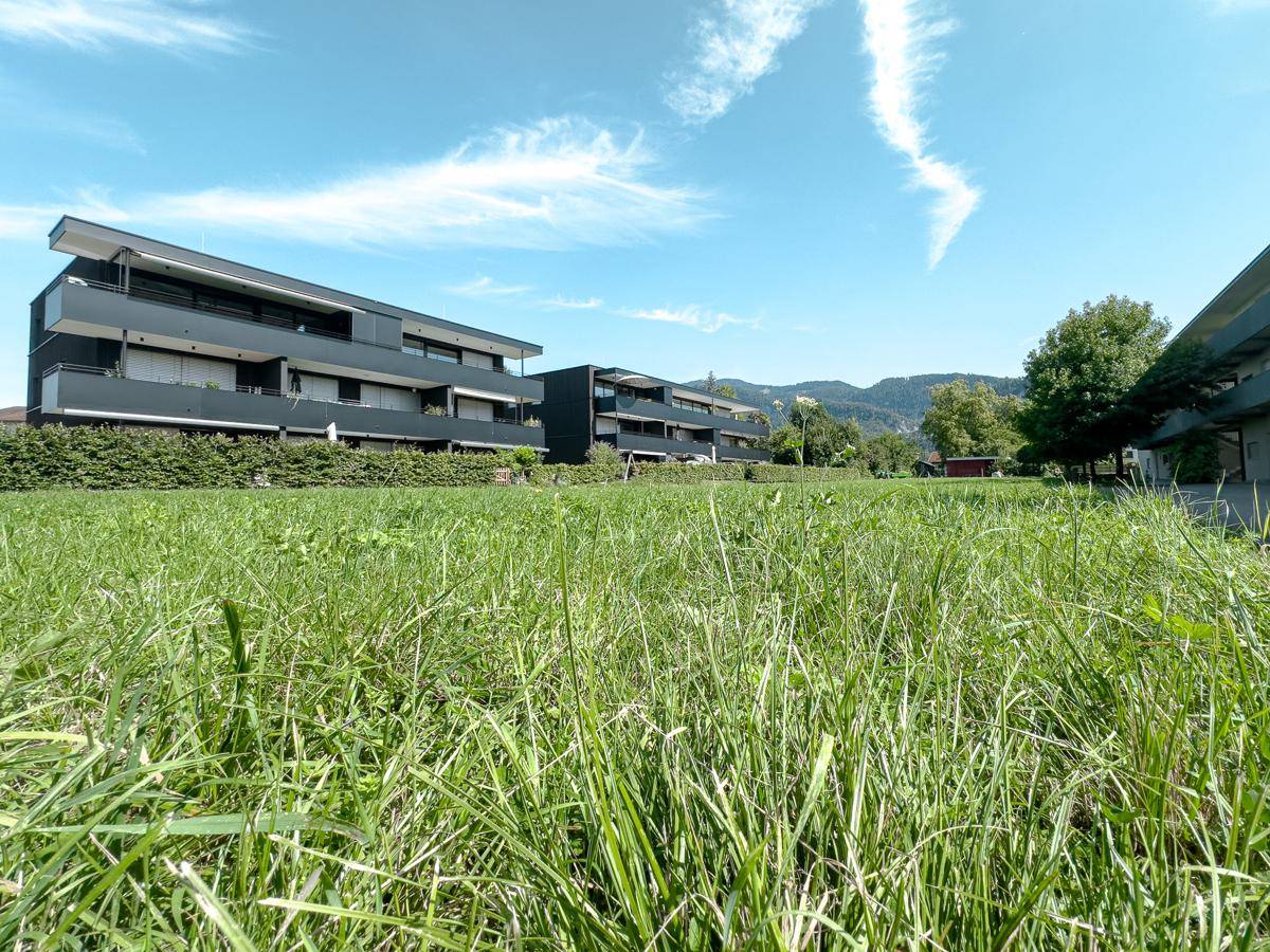 Blick über eine weite Rasenfläche auf moderne Wohngebäude und die umliegende Berglandschaft.