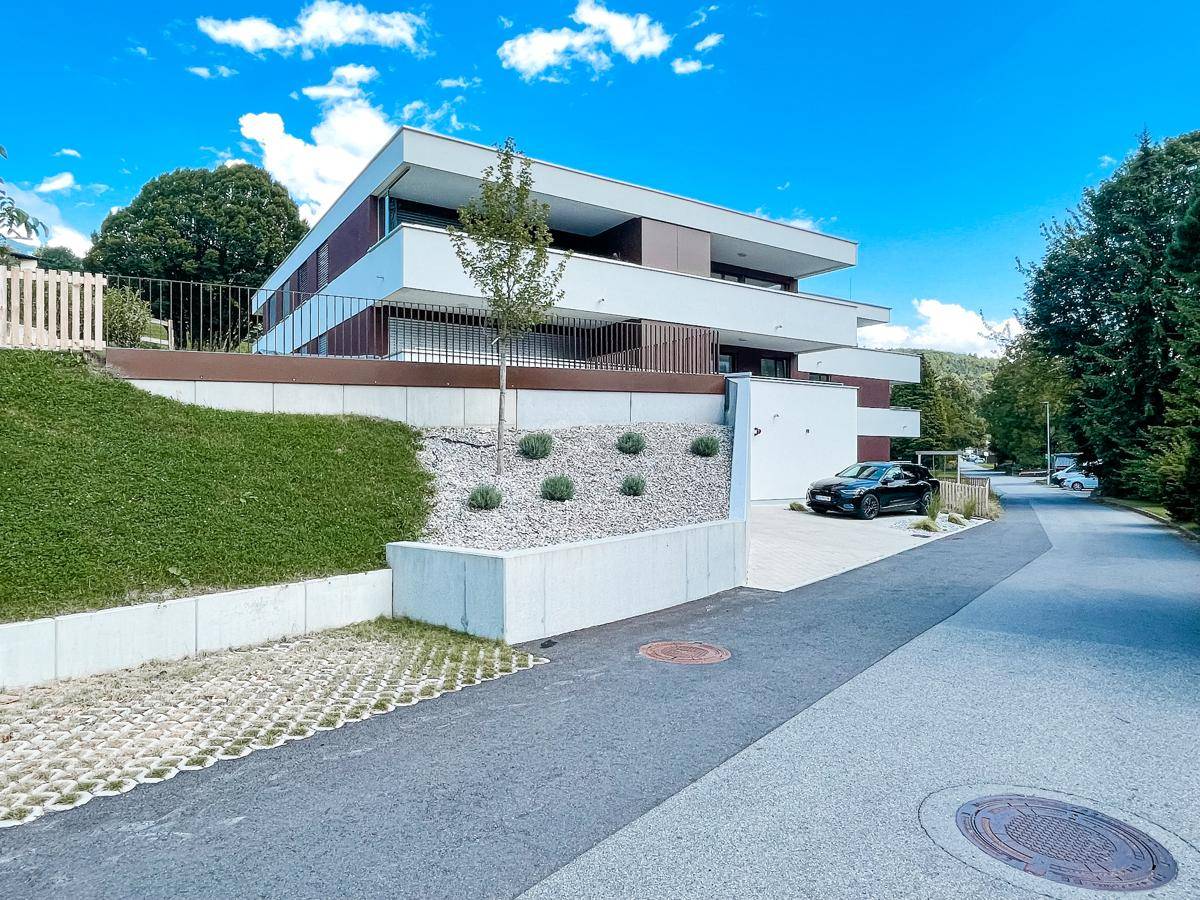Modernes Mehrfamilienhaus mit mehreren Balkonen, umgeben von Grünflächen und einer Zufahrt.