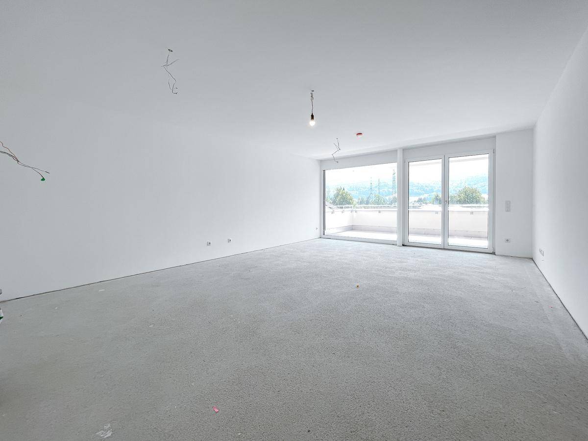 Heller, unfertiger Wohnraum mit großen Fenstern und Zugang zu einem Balkon mit Bergblick.