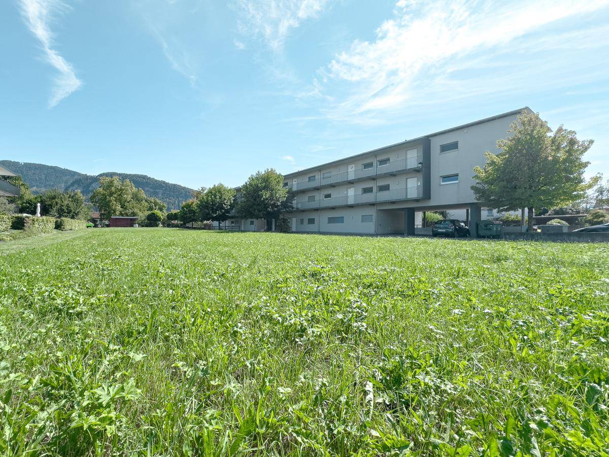 Weitläufige Rasenfläche vor einem Mehrfamilienhaus mit Balkonen und Bergkulisse.