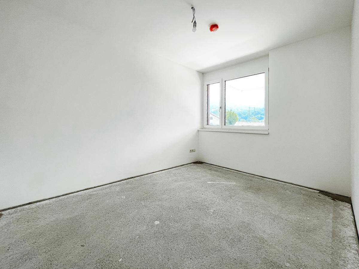 Unfertiges Zimmer mit weiß gestrichenen Wänden, Betonboden und einem Fenster mit Blick ins Grüne.