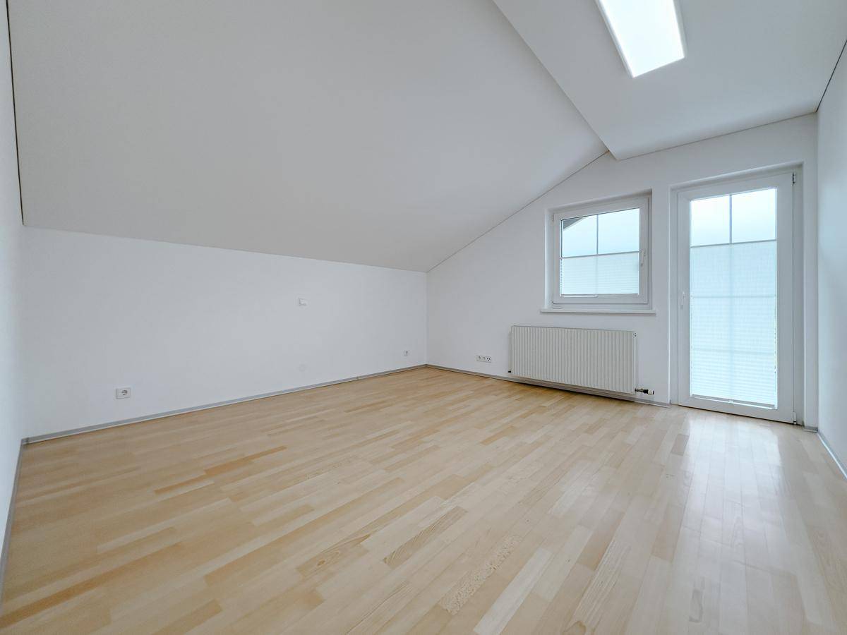 Geräumiges Zimmer im Dachgeschoss mit Parkettboden, Fenster und Zugang zu einem Balkon.