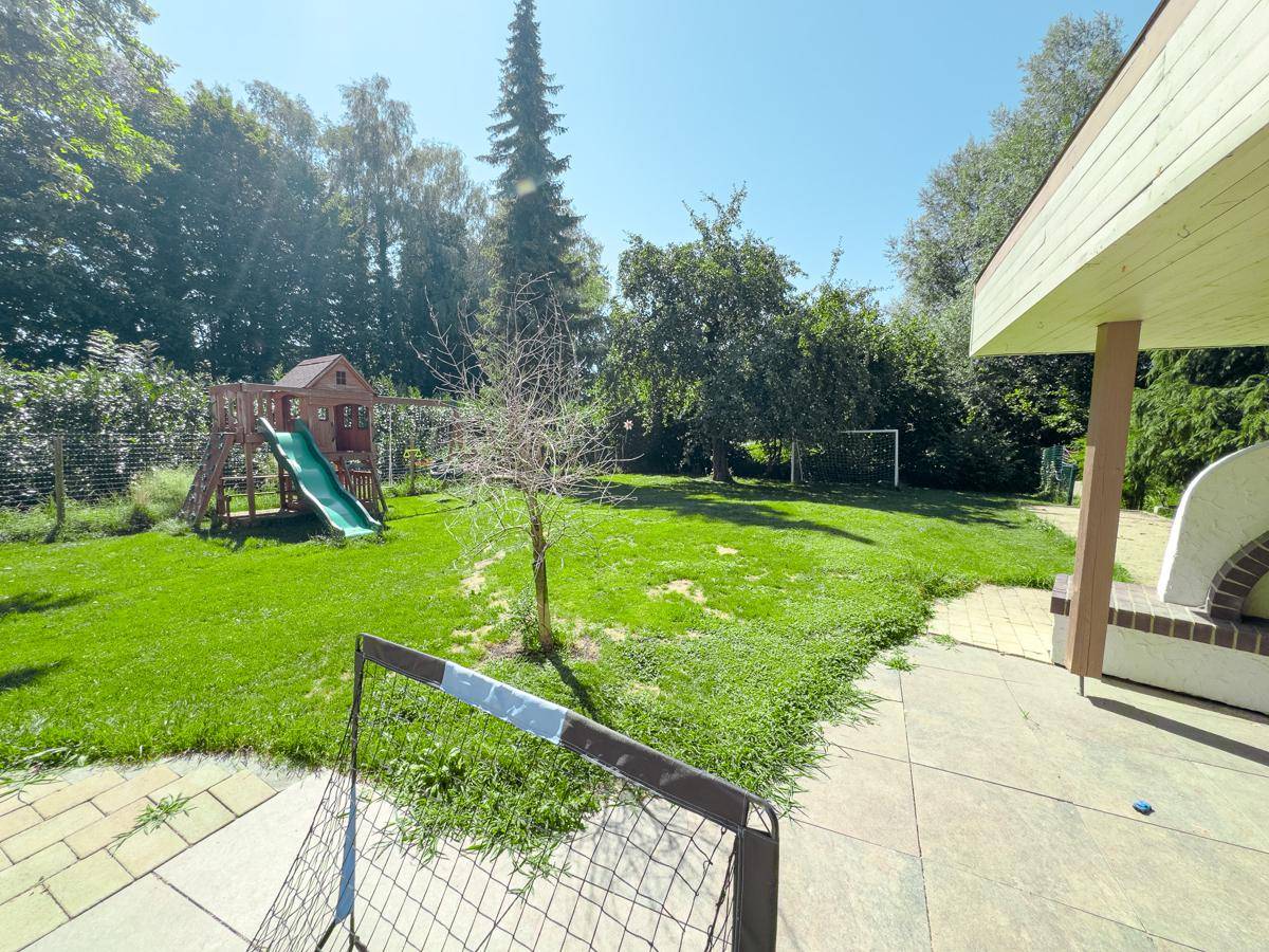 Terrasse mit Blick auf den grünen Garten, Spielplatz, Fußballtor und einen gemauerten Außenkamin.