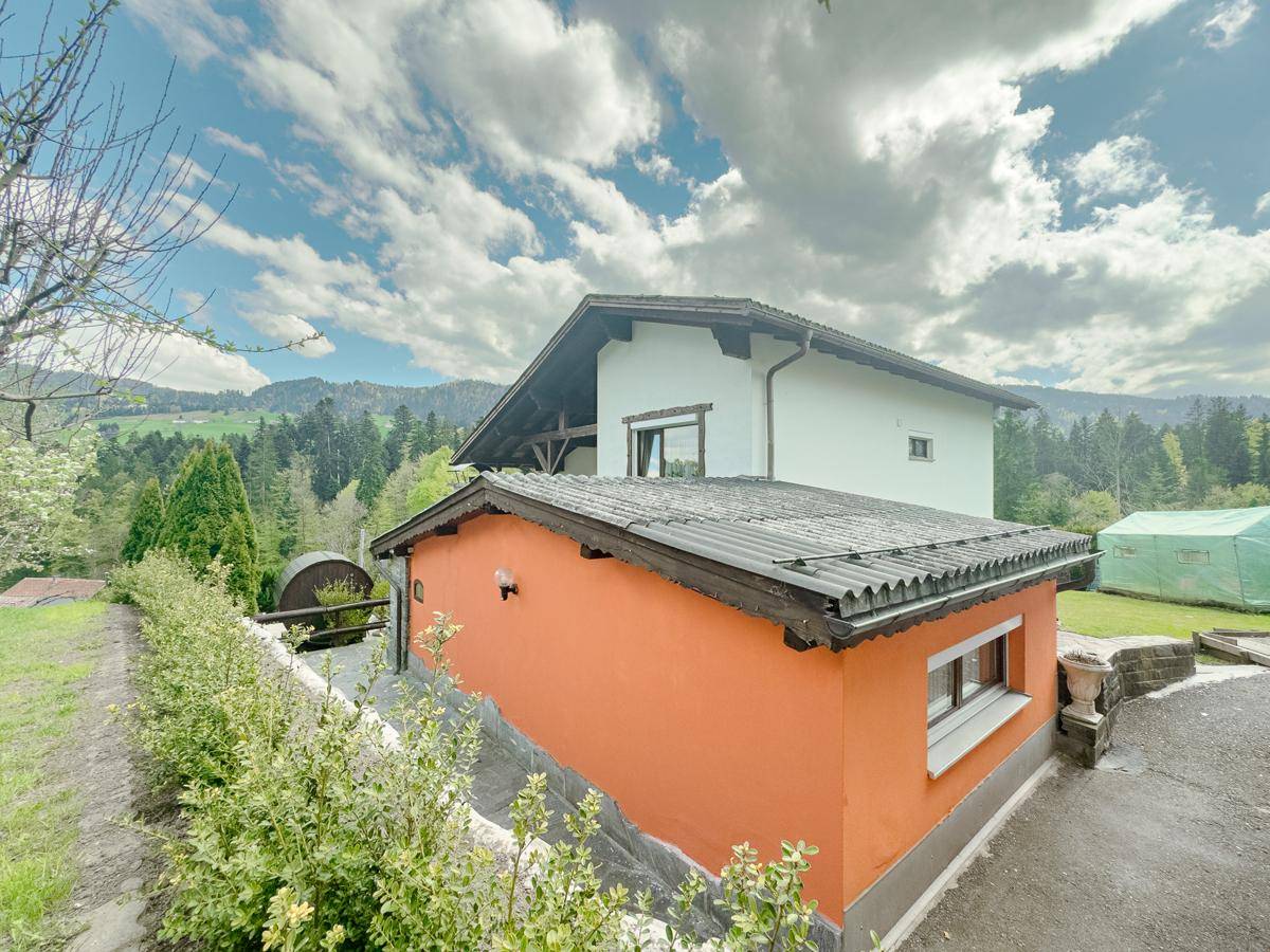 Seitenansicht des Hauses mit orangefarbenem Sockel und weißer Fassade vor Bergkulisse.