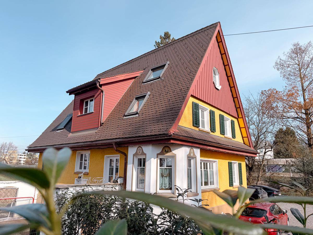 Charmantes Haus mit gelber Fassade, rotem Dach und Dachfenstern, umgeben von üppigem Grün.