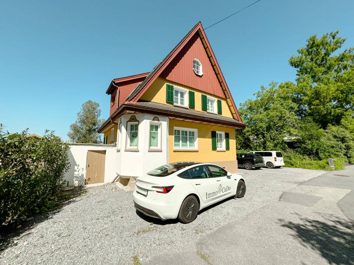 Traditionelles Haus mit gelber Fassade, rotem Dach und Schotterauffahrt, umgeben von viel Grün.