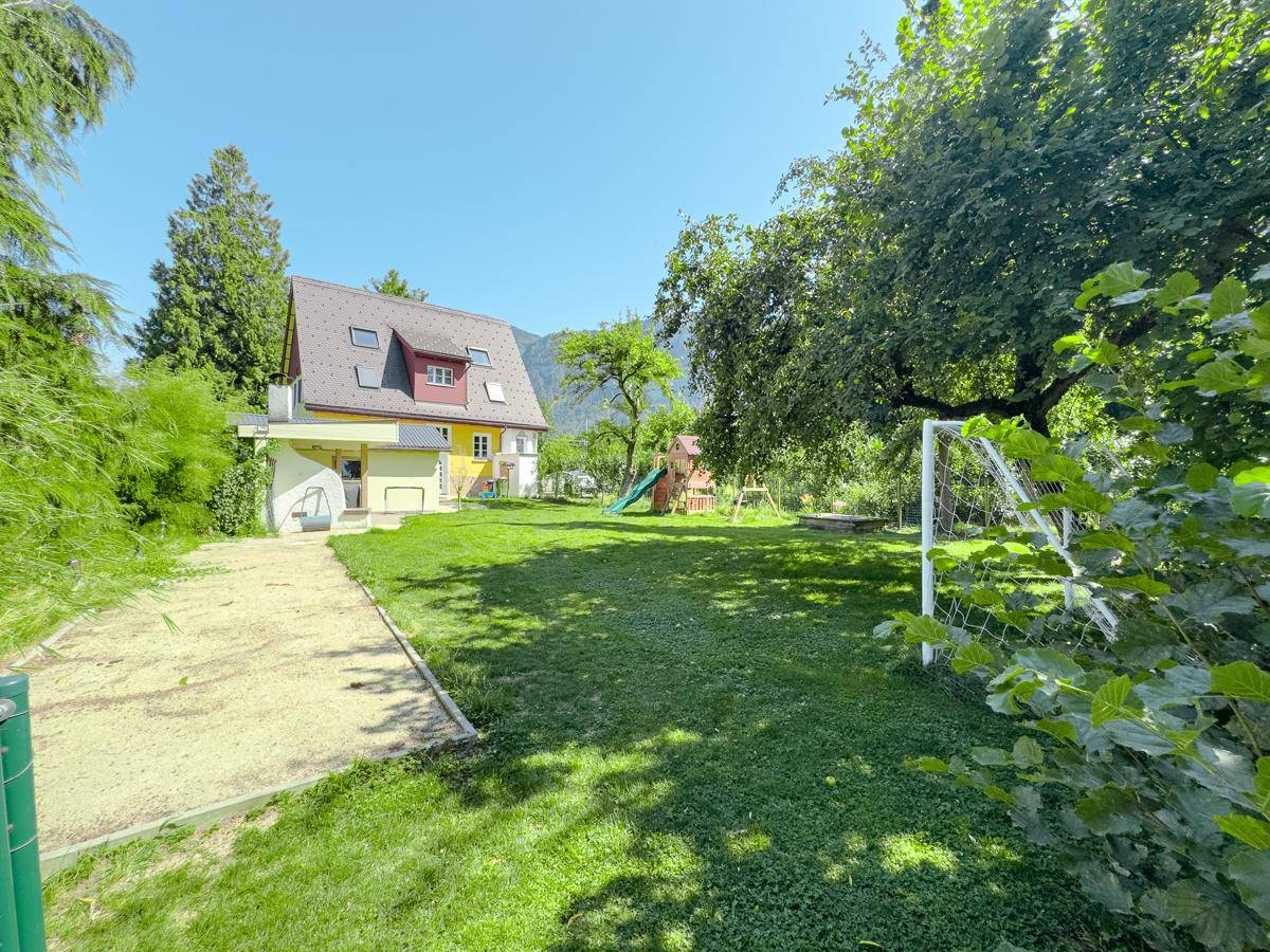 Großer Garten mit Haus, Spielplatz, Rasenfläche und einem kleinen Fußballtor vor Bergkulisse.
