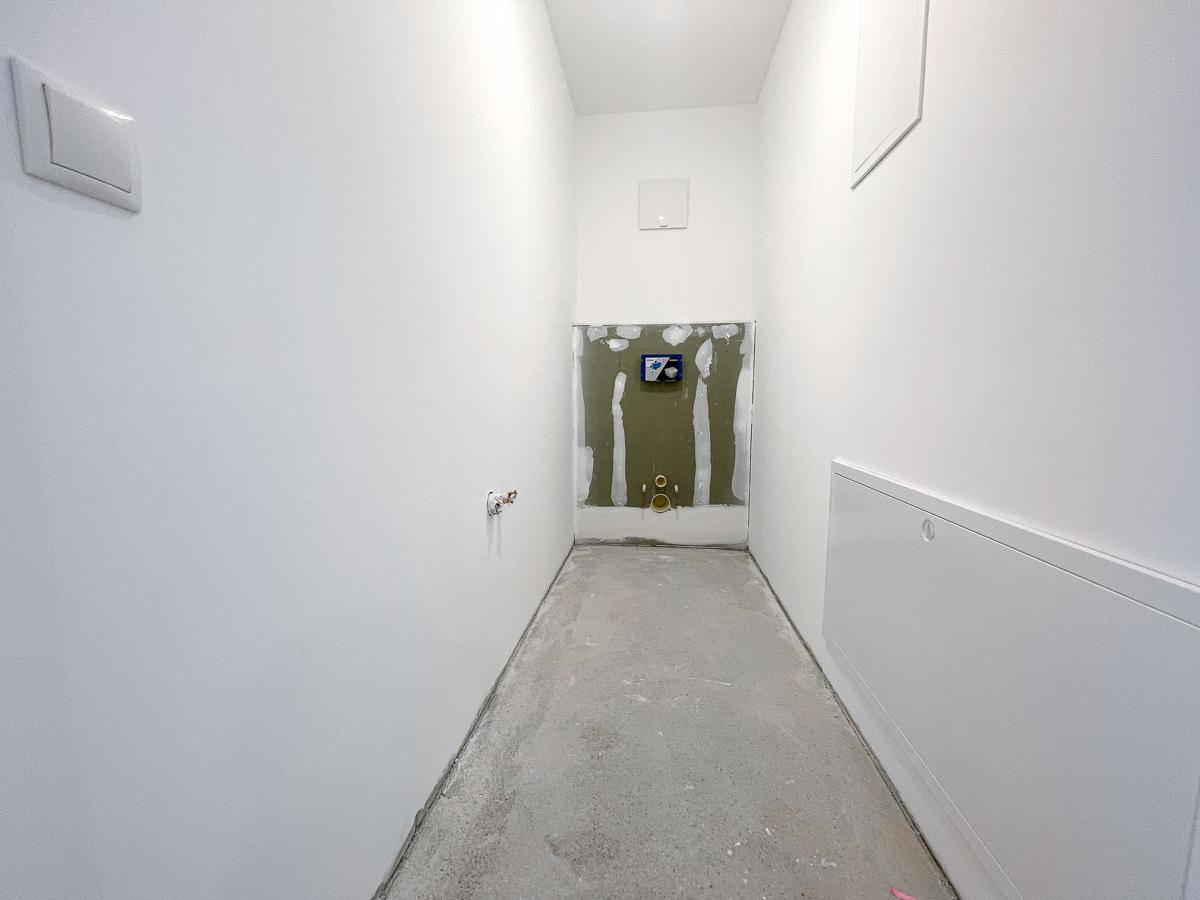 Schmaler Raum im Bau, vorbereitet für ein WC, mit sichtbaren Installationen und Betonboden.