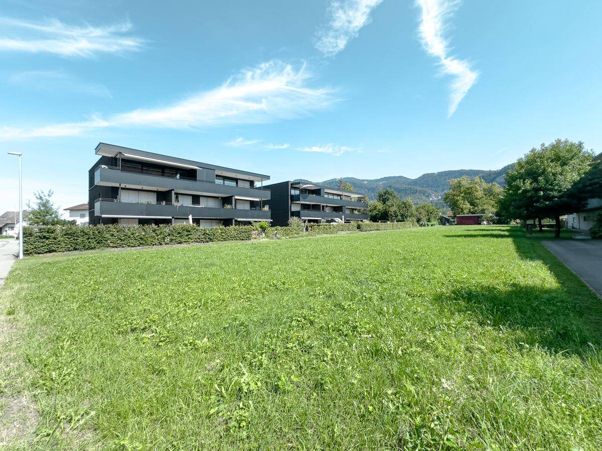 Blick über eine grüne Wiese auf moderne Wohngebäude mit Balkonen und die umliegende Natur.