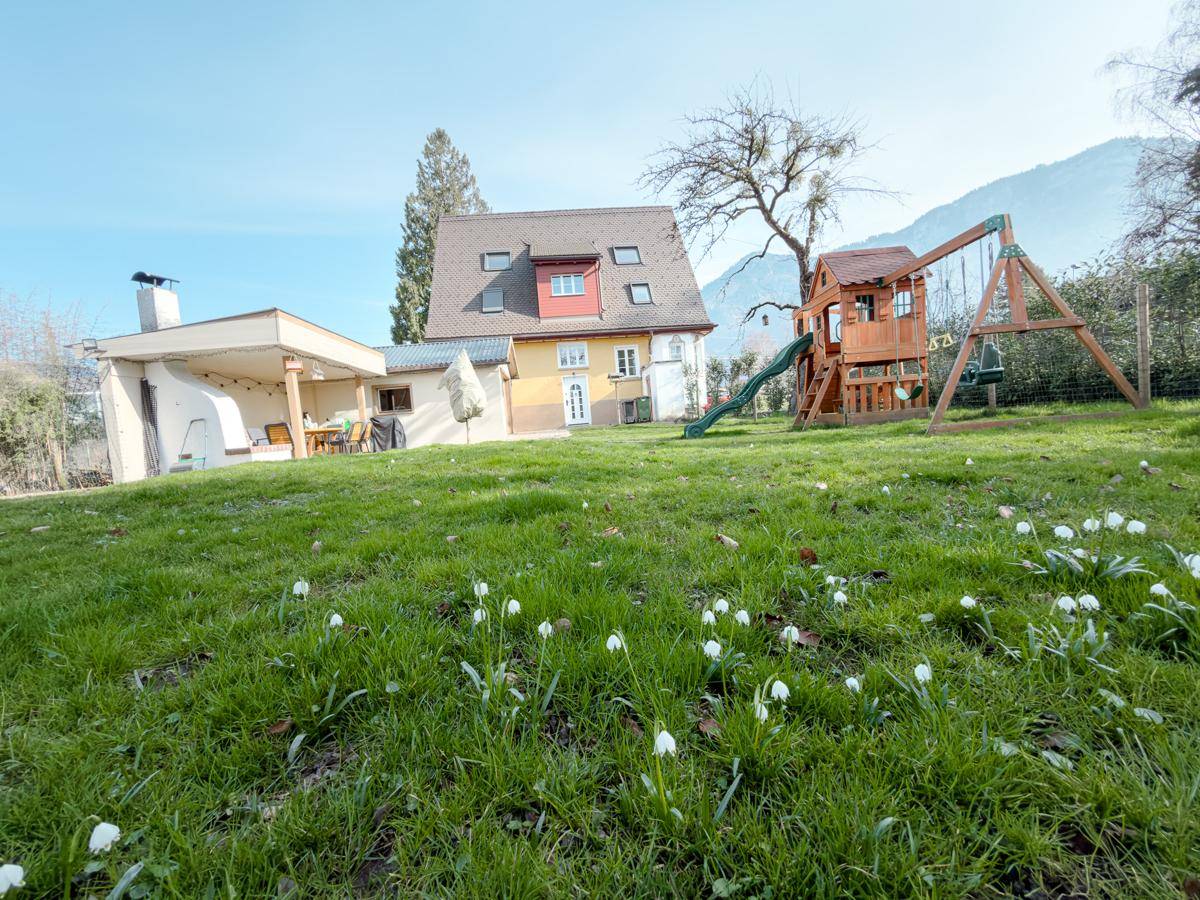 Großer, gepflegter Garten mit Spielplatz, Rasenfläche und Blick auf die Berge im Hintergrund.
