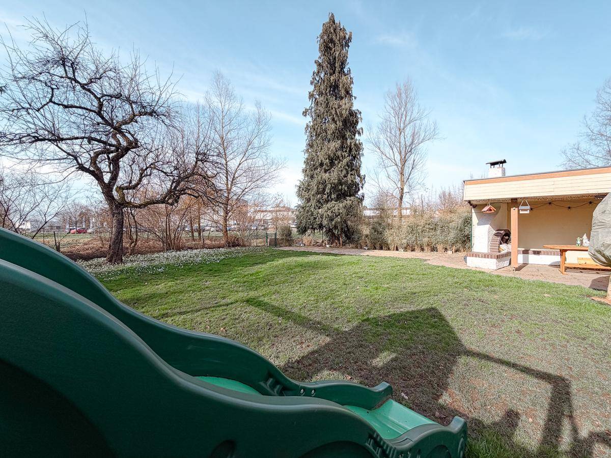 Grüner Garten mit Spielplatzrutsche und Blick auf den überdachten Grillbereich im Hintergrund.