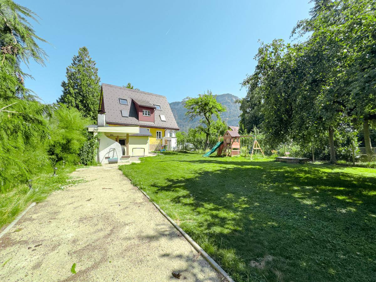 Weitläufiger Garten mit großem Rasen, Spielplatz und Blick auf das Haus sowie die umliegenden Berge.