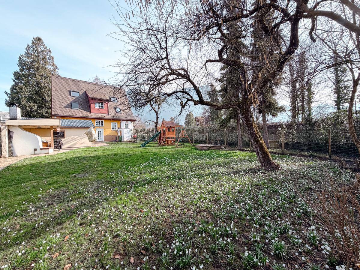 Weitläufiger Garten mit Spielplatz, Rasen und blühenden Schneeglöckchen, umgeben von Bäumen.