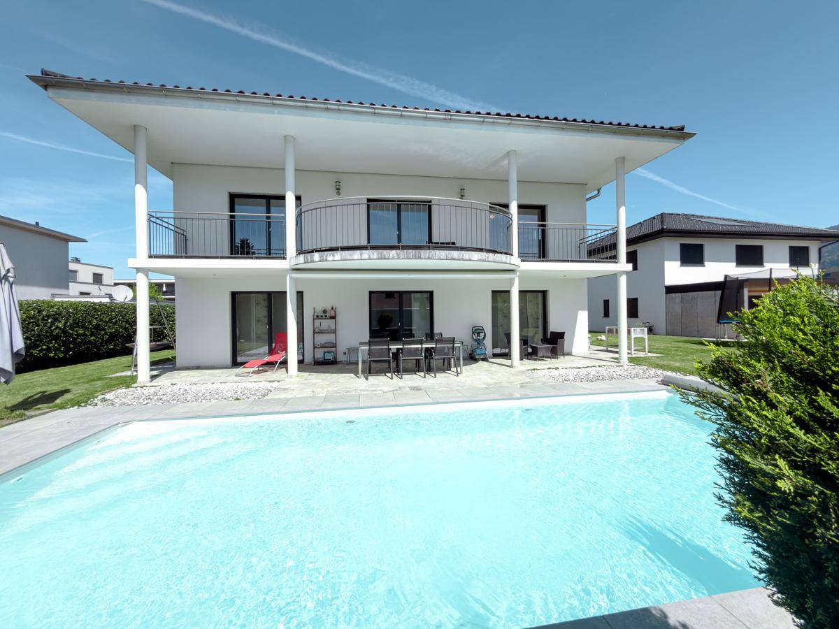 Rückansicht des Hauses mit großem Pool und möblierter Terrasse im Sonnenschein.