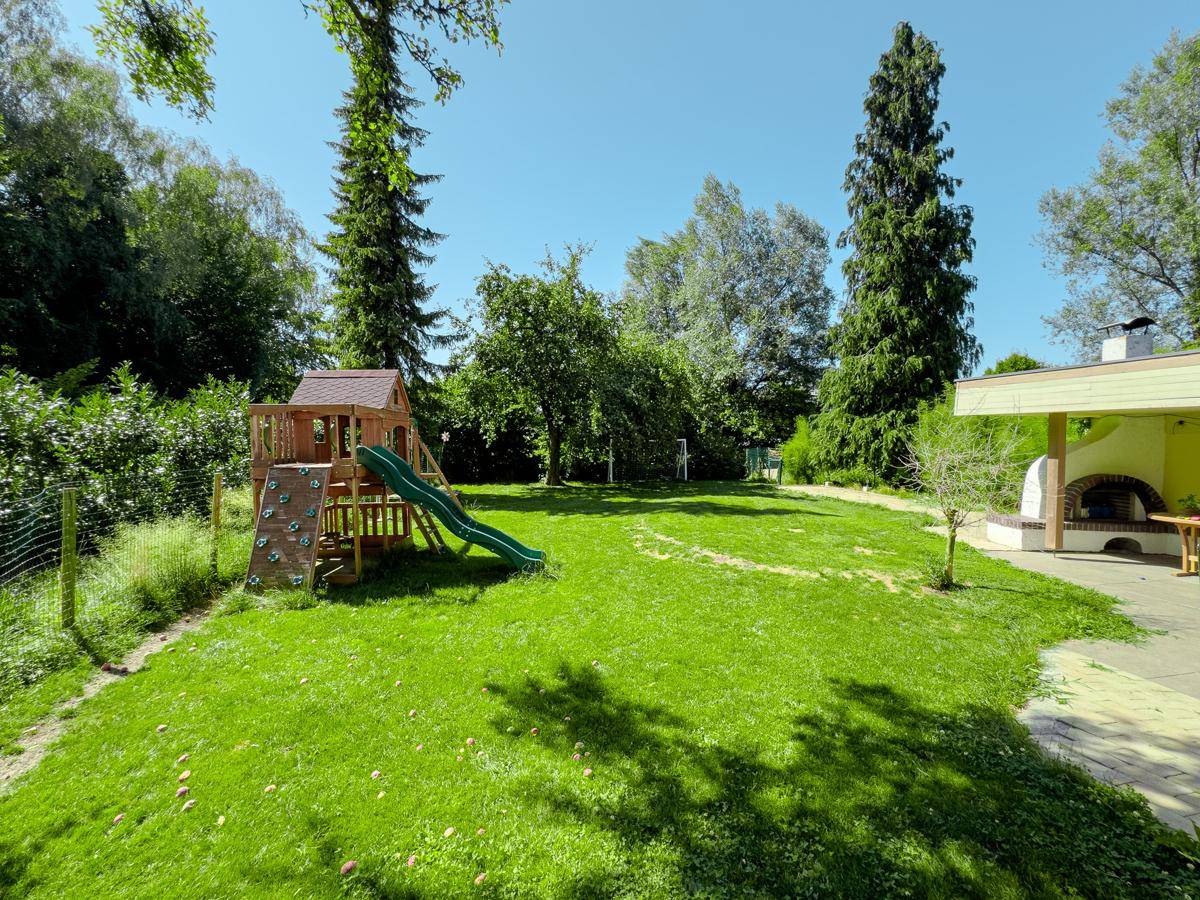 Grüner Garten mit Spielplatz, Rasenfläche und einem gemauerten Pizzaofen neben dem überdachten Sitzbereich.