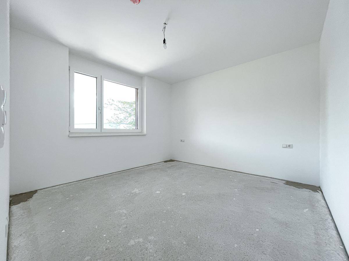 Leerer Raum im Rohbau mit Fenster, weißen Wänden und Estrichboden, wartet auf den Innenausbau.