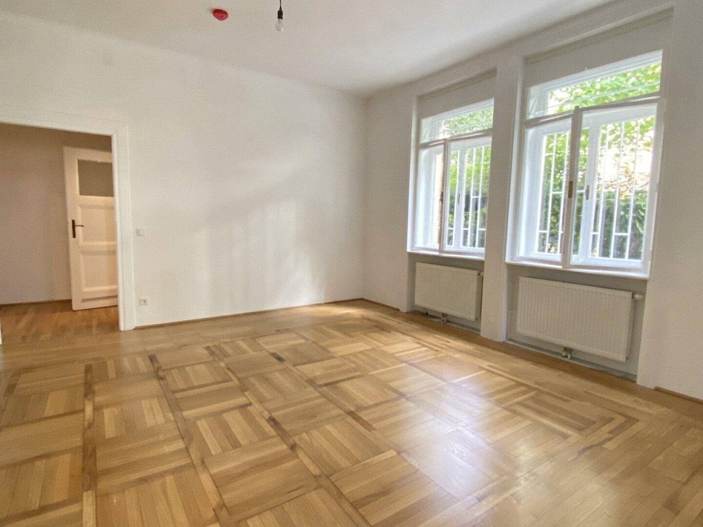 Geräumiges Zimmer mit hellem Parkettboden und zwei großen Fenstern, die viel Tageslicht hereinlassen und eine grüne Aussicht bieten.