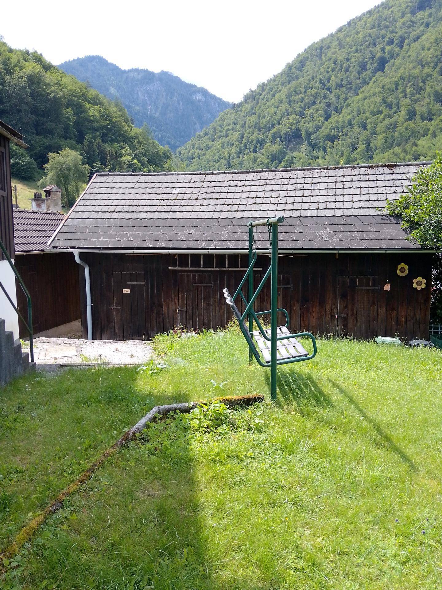 Grüner Gartenbereich mit Schaukel und rustikalem Holzschuppen vor Bergkulisse.