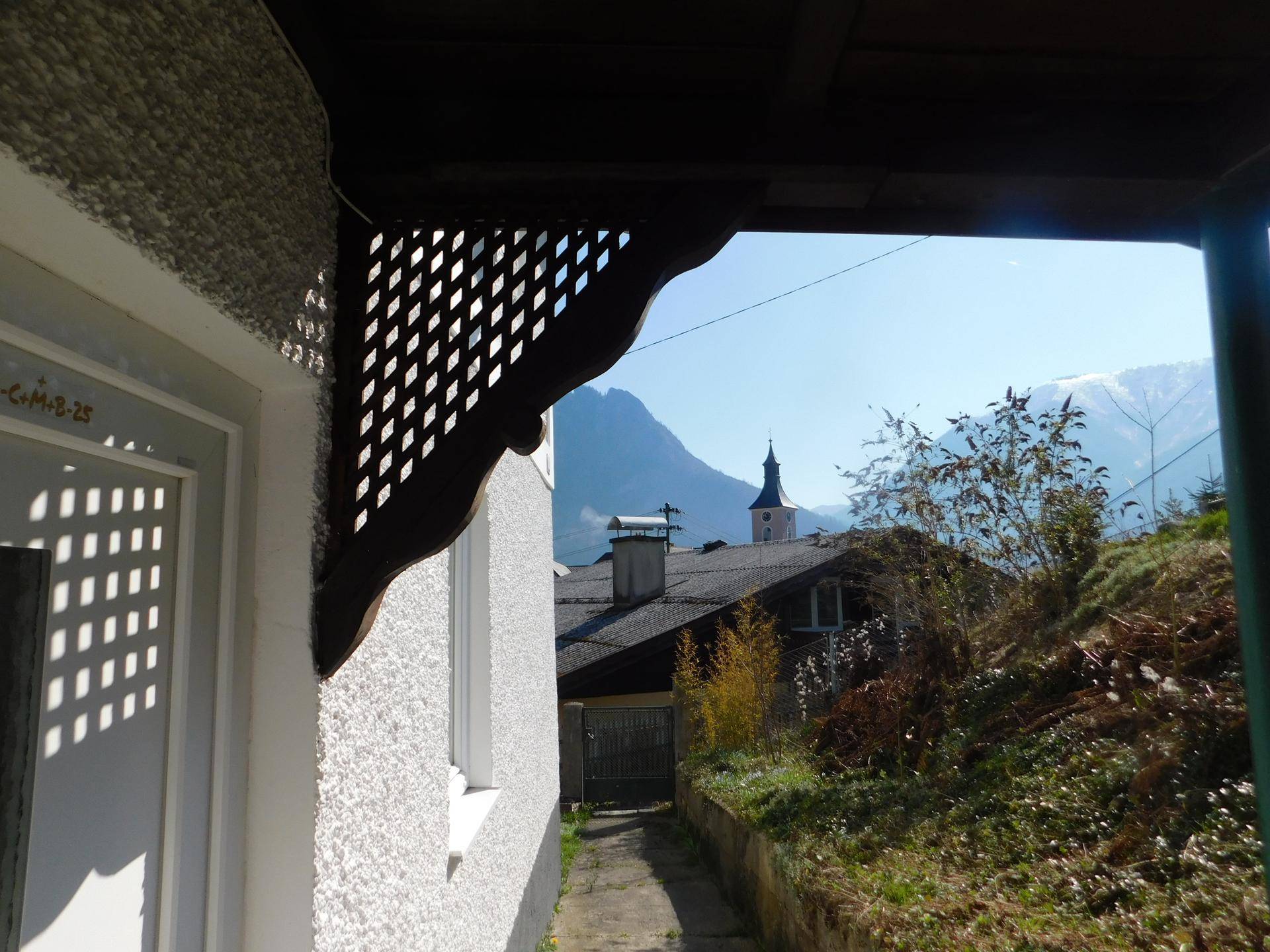 Blick unter einem Vordach hindurch auf einen schmalen Weg, Dächer und Berge mit Kirchturm.