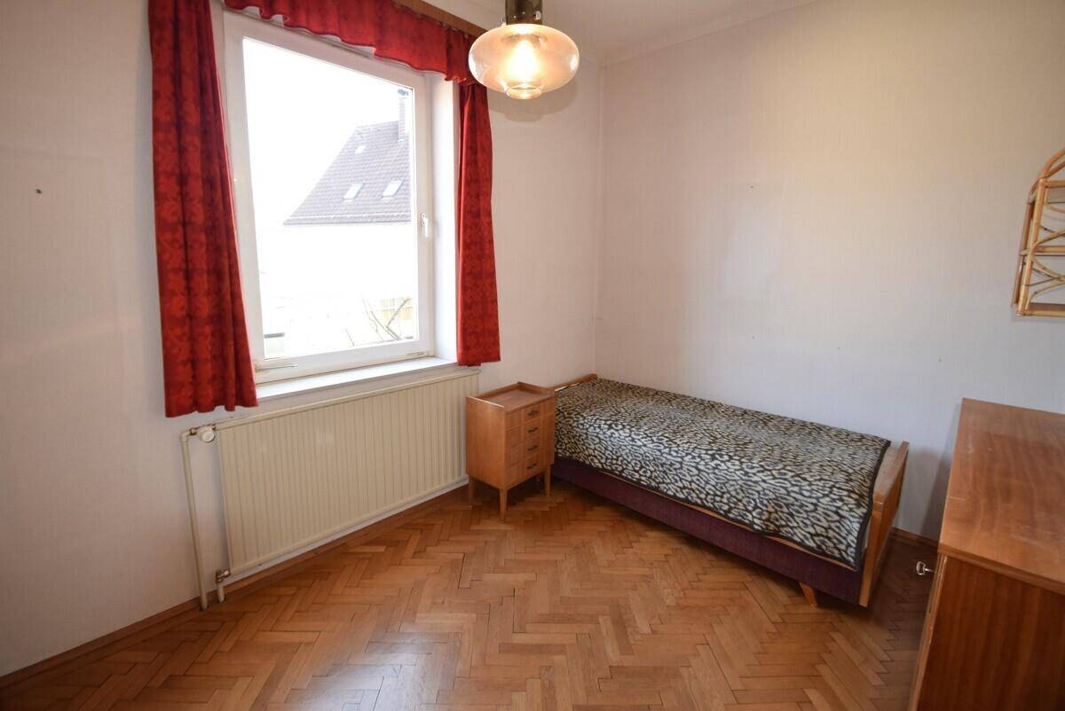 Schlafzimmer mit Parkettboden, großem Fenster und einfacher Einrichtung.