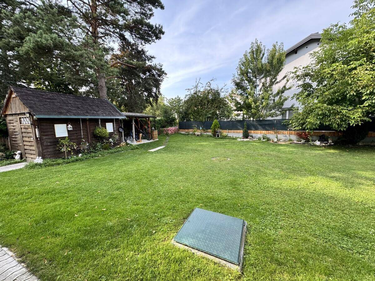 Großer, gepflegter Garten mit Rasenfläche, einem Holzschuppen und hohen Bäumen im Hintergrund.