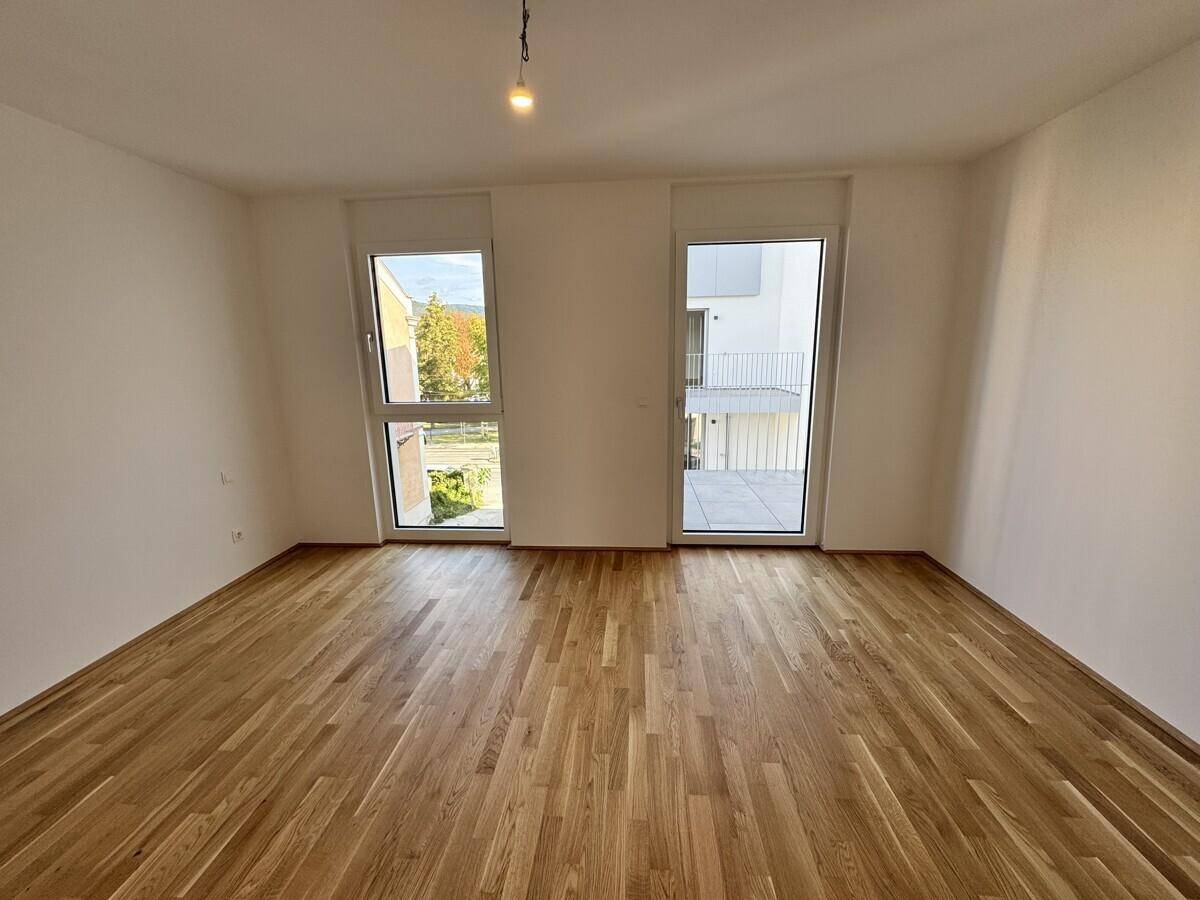 Leerer Raum mit Holzboden und zwei hohen Fenstern, die viel Tageslicht hereinlassen.