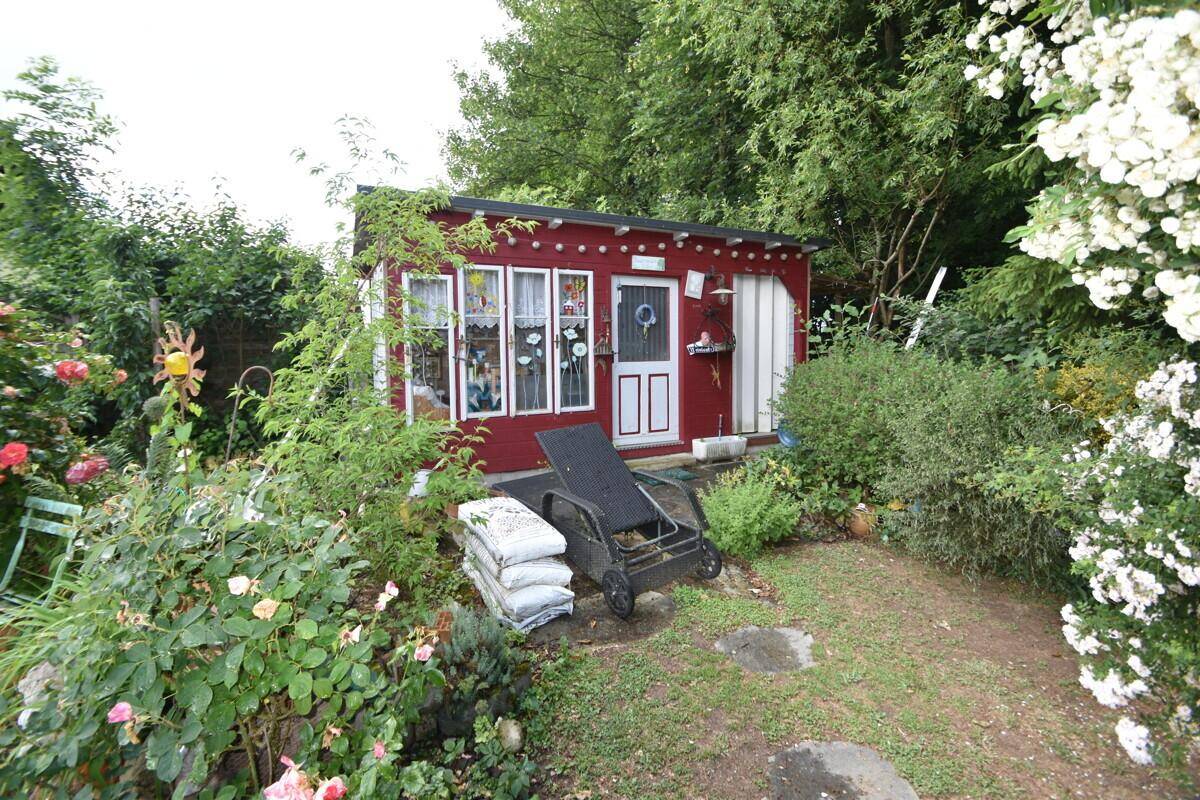 Kleines, rotes Gartenhaus umgeben von üppiger Vegetation und Blumen.