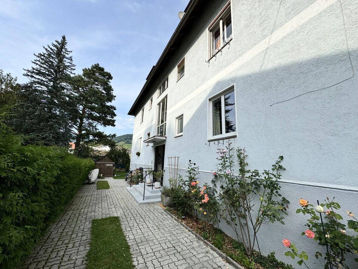 Seitenansicht des Gebäudes mit gepflastertem Weg, Rosensträuchern und grüner Umgebung.