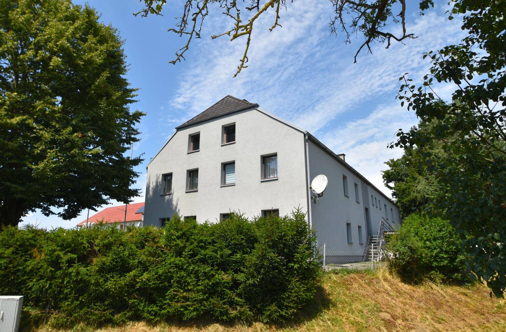 Rückansicht eines Mehrfamilienhauses mit Satellitenschüssel, grüner Hecke und Bäumen.