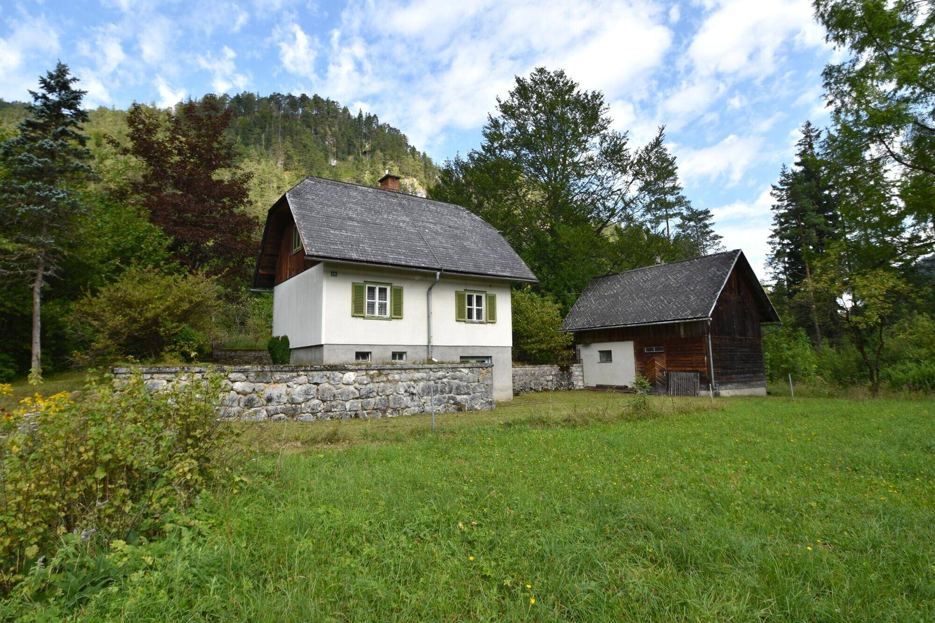 Traditionelles Haus mit Holzgiebel und Nebengebäude, umgeben von einer grünen Wiese und Bäumen.