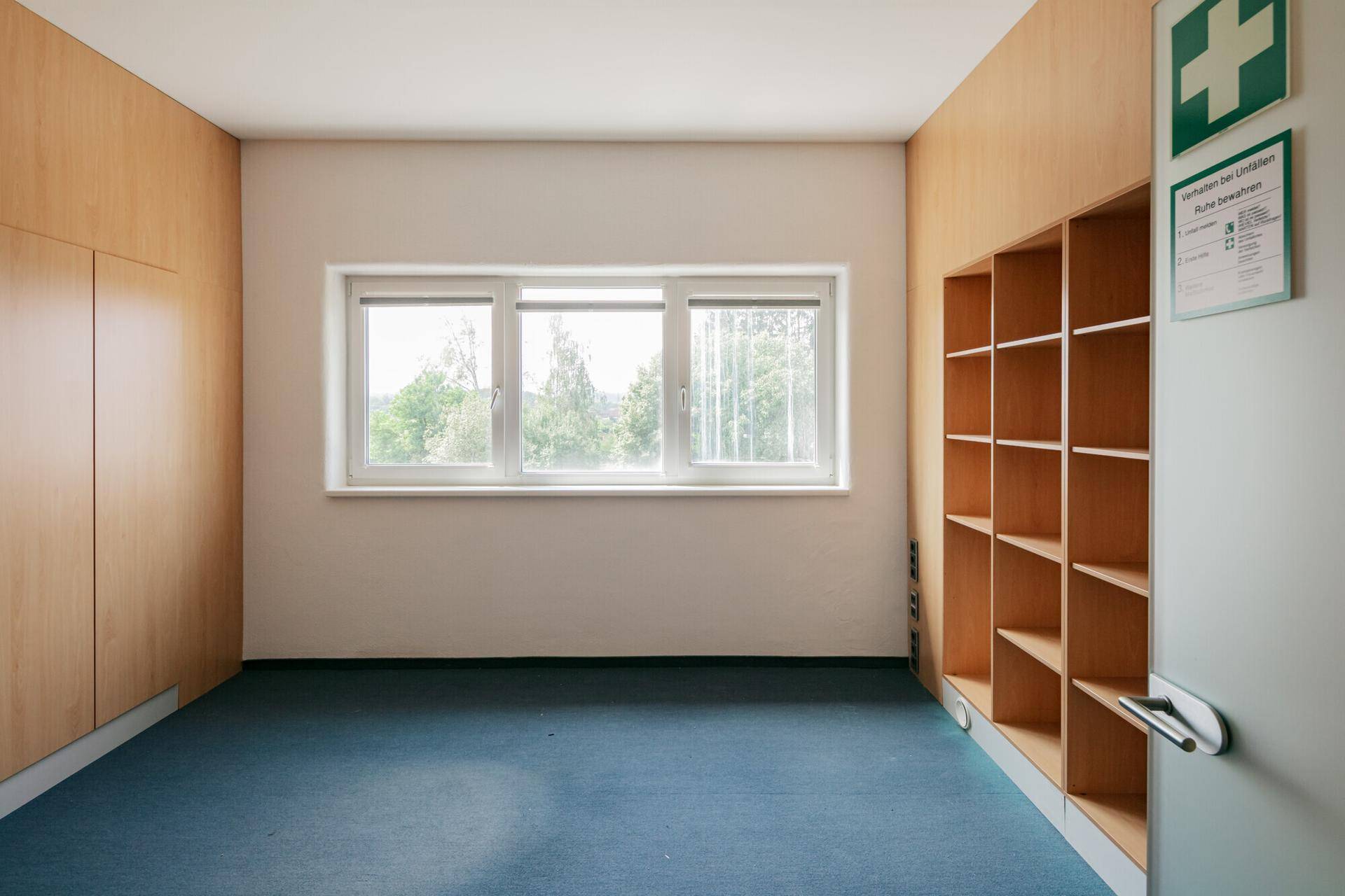 Kleiner Raum mit blauem Teppichboden, weißen Wänden, Holzschränken und einem Fenster mit Blick ins Grüne.