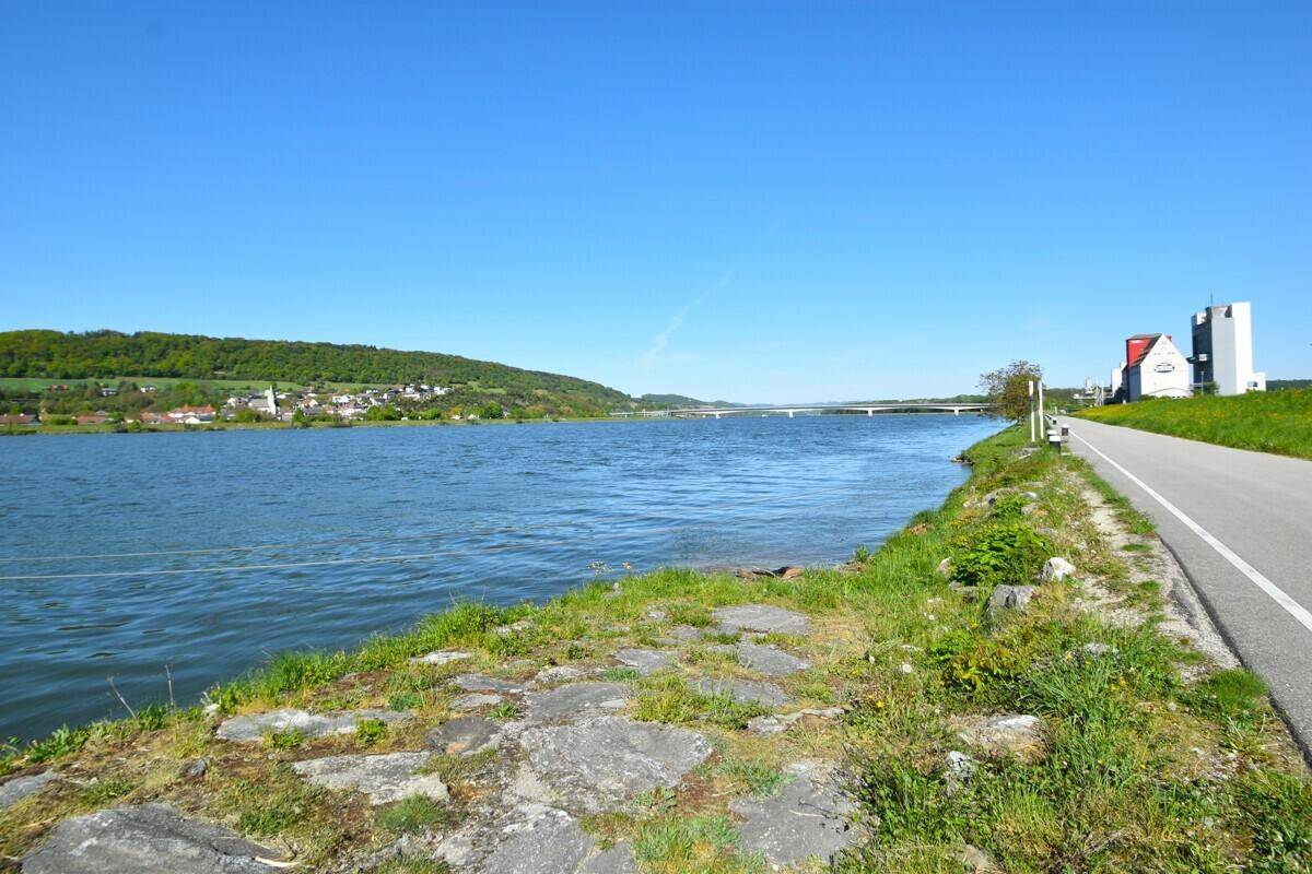 Weitläufiger Fluss mit Uferstraße, grünen Hügeln und einer Brücke in der Ferne unter blauem Himmel.