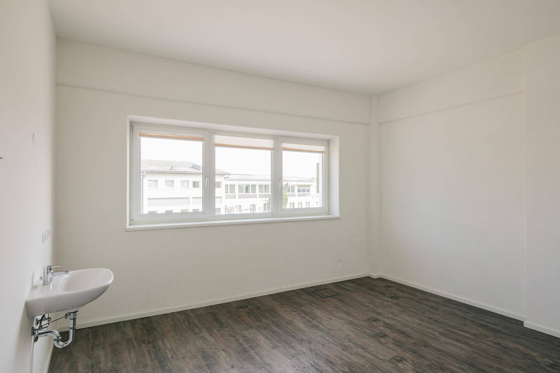 Heller Büroraum mit großem Fenster, Holzoptik-Boden und einem integrierten Waschbecken.