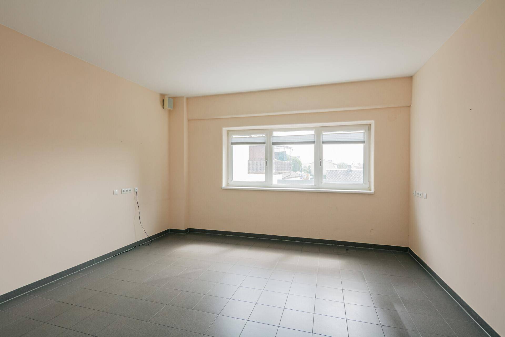 Leerer Raum mit beigefarbenen Wänden, grauen Fliesen und einem großen Fenster, das Tageslicht hereinlässt.
