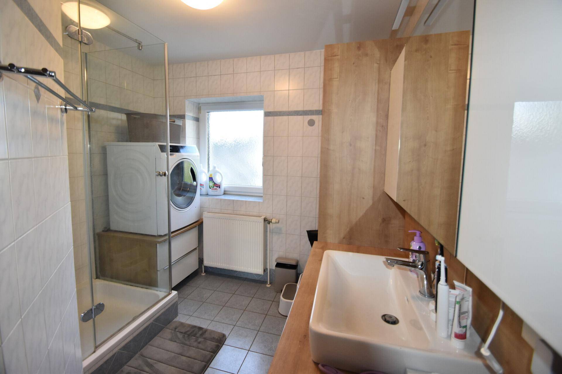 Gefliestes Badezimmer mit Dusche, Waschbecken und Waschmaschine unter einem Fenster.