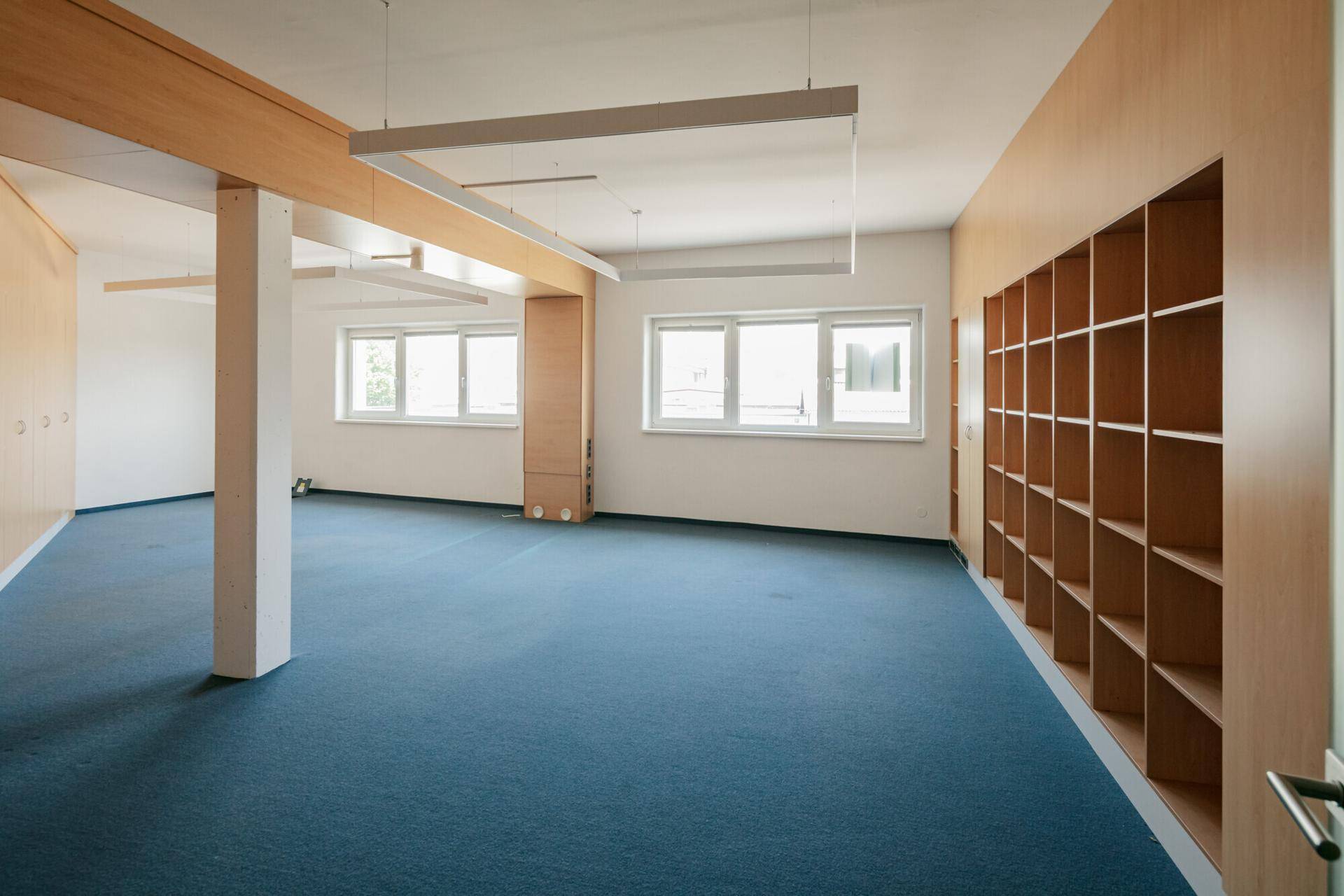 Großes, leeres Büro mit blauem Teppichboden, Holzverkleidungen und integrierten Regalen entlang der Wand.