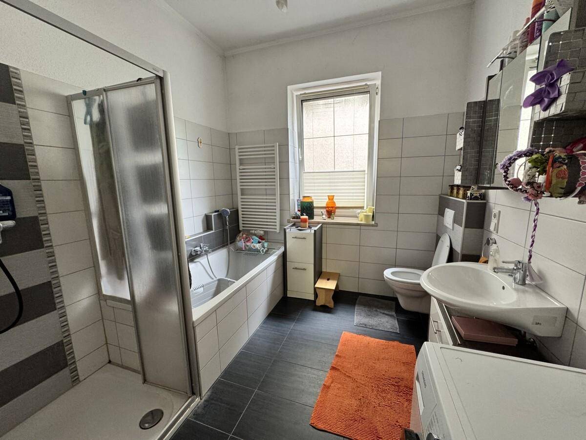 Badezimmer mit Badewanne, Dusche, Fenster und weiß-grauen Fliesen.
