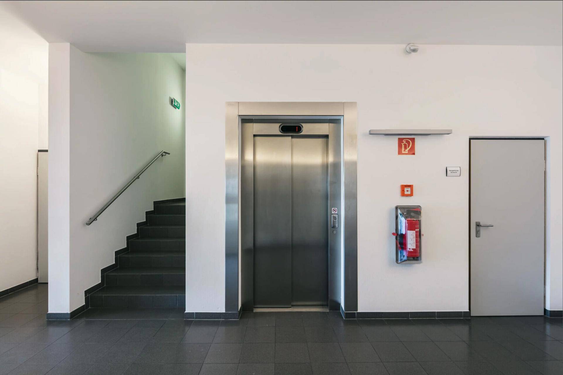 Heller Flur mit modernem Aufzug, Treppe und dunklen Fliesenböden im Eingangsbereich.
