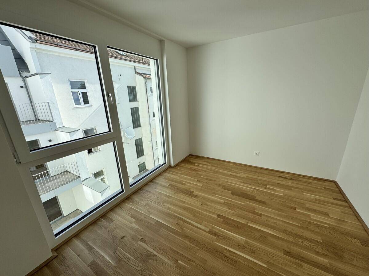 Leeres Zimmer mit großen Fenstern und Holzboden, bietet einen Blick auf den Innenhof des Gebäudes.