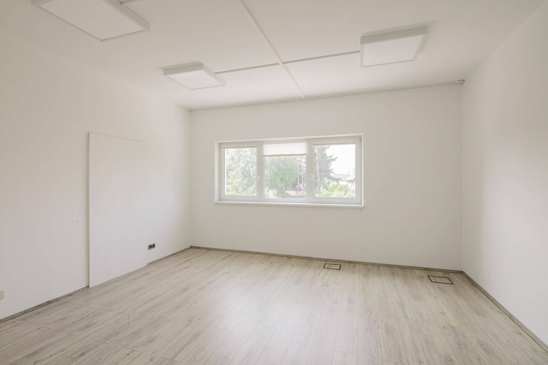Heller Büroraum mit großem Fenster, hellem Laminatboden und weißen Wänden.