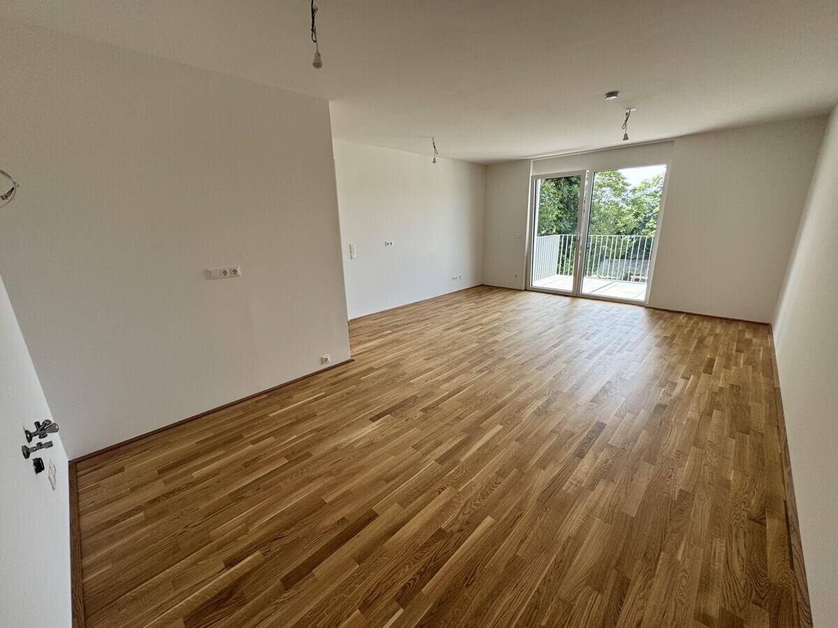 Heller, leerer Raum mit Eichenparkettboden und Zugang zu einem Balkon mit grüner Aussicht.
