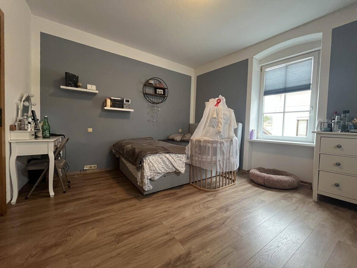 Schlafzimmer mit Doppelbett, Babybett und Schminktisch, akzentuiert durch eine graue Wand.