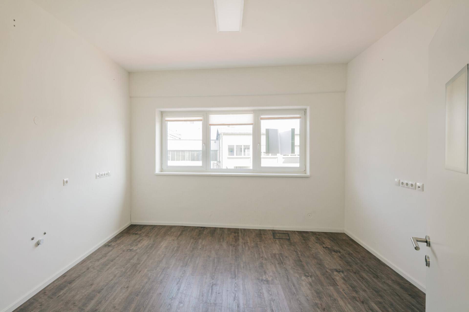 Leerer Büroraum mit großen Fenstern, die viel Tageslicht hereinlassen, und dunklem Holzoptik-Boden.