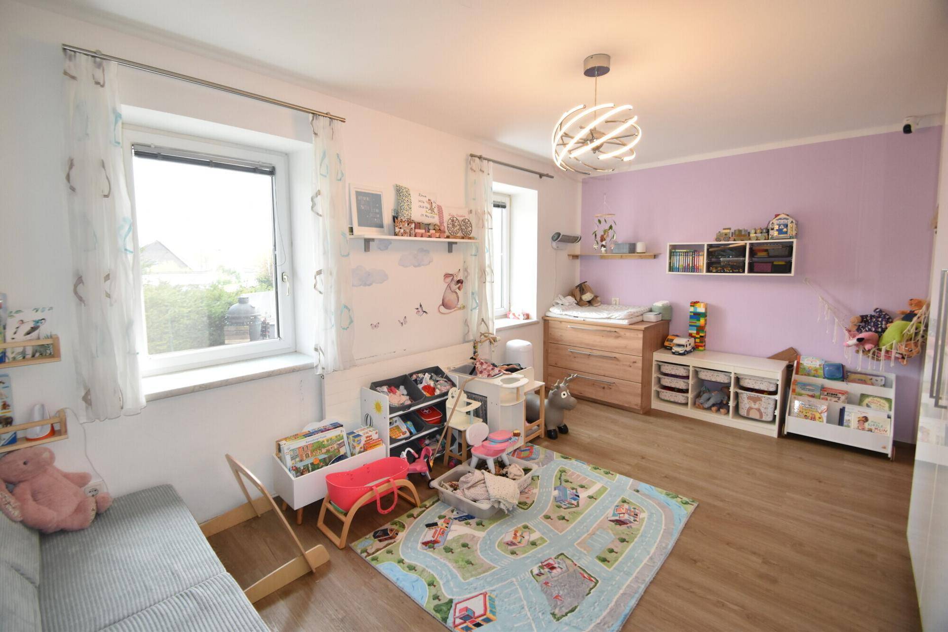 Helles Kinderzimmer mit Spielteppich, Wickelkommode und lila Akzentwand.