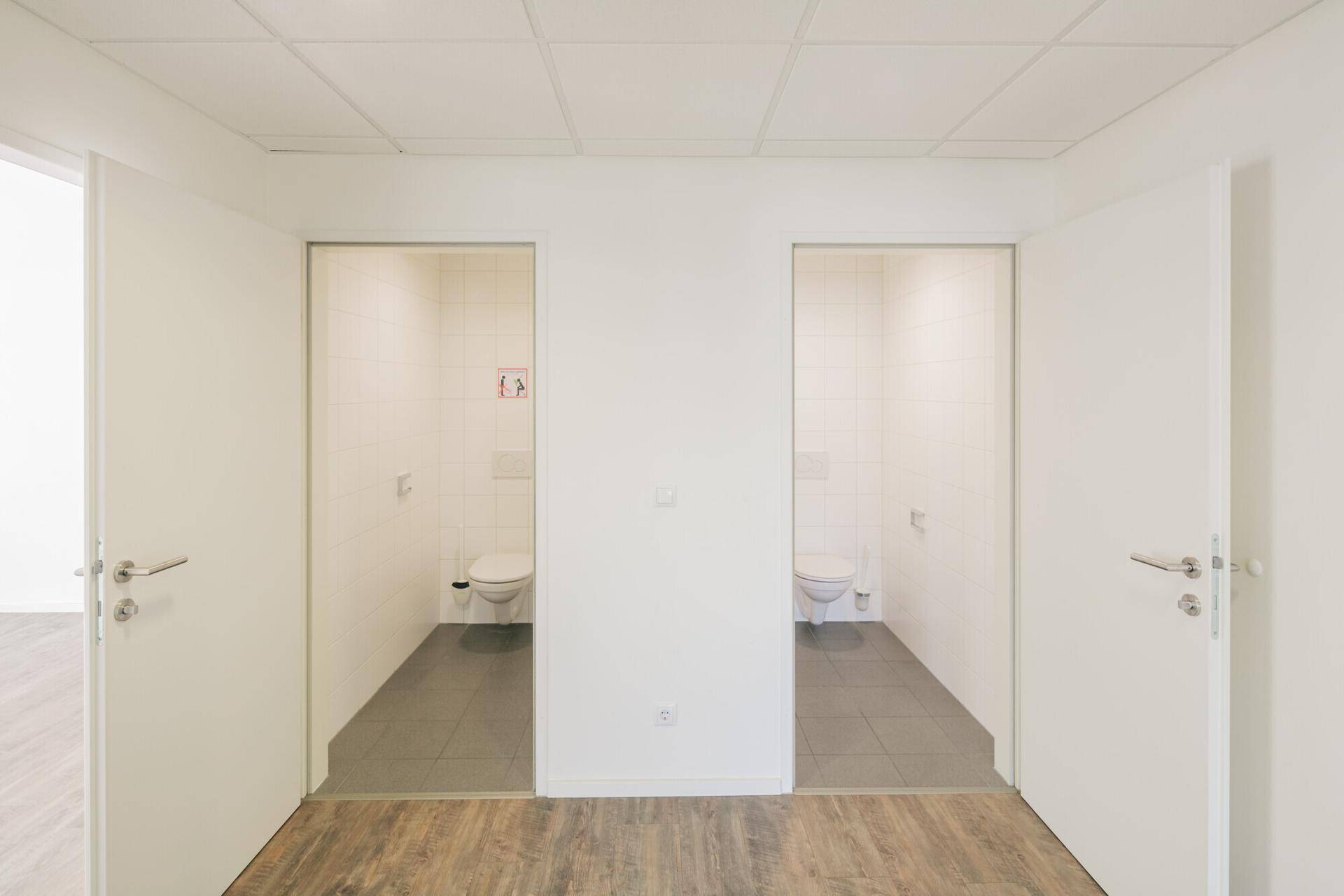 Zwei separate Toilettenräume mit weißen Fliesen und Wand-WCs, zugänglich von einem gemeinsamen Flur.
