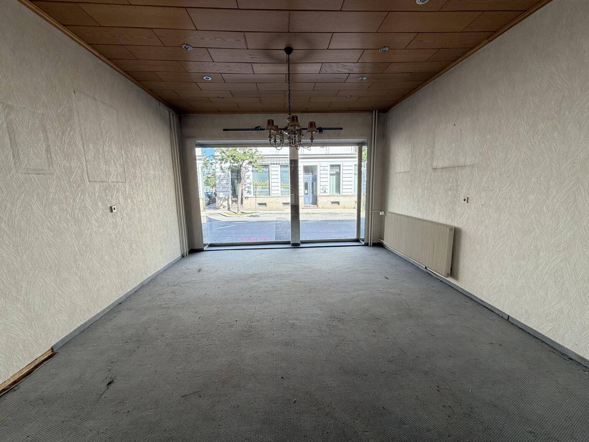 Großer, leerer Raum mit Teppichboden, Holzdecke und großen Fenstern zur Straße.