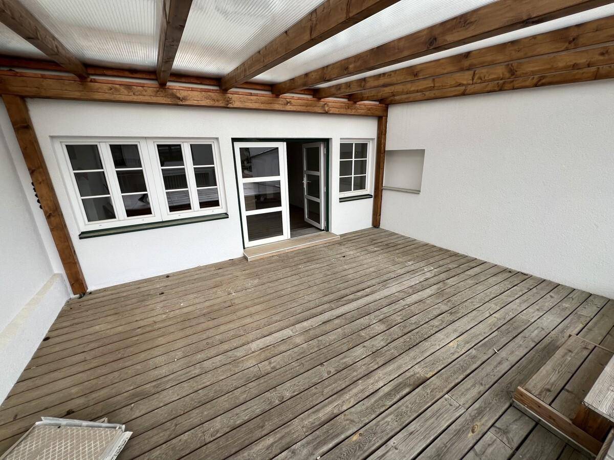 Überdachte Holzterrasse mit Holzbalken und Zugang zu Innenräumen durch große Fenster.
